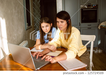 Mother and daughter using laptop sitting together in home kitchen. Mom helping kid with school homework. Education with modern technologies. Mother and daughter using laptop sitting together in home kitchen. Mom helping kid with school homework. Education with modern technologies. 125724589