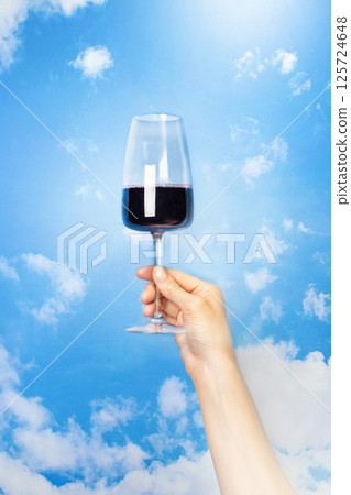 A woman's hand holds a glass of red wine against a blue sky with clouds, vertical banner A woman's hand holds a glass of red wine against a blue sky with clouds, vertical banner 125724648