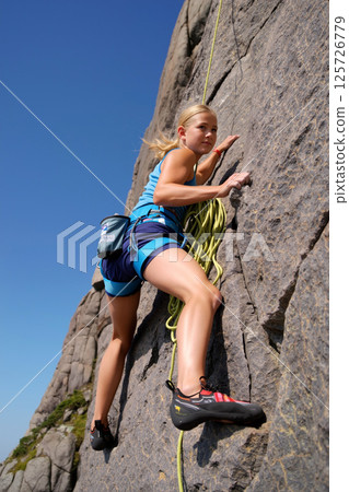 Young Climber Ascends a Rocky Cliff Against a Clear Blue Sky on a Sunny Day in Nature 125726779