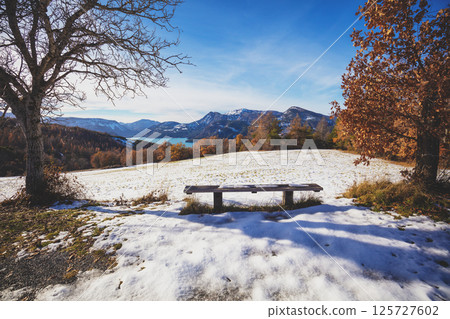 Wooden bench near the mountain lake in winter on a sunny day. View of Serre-Poncon mountain lake in winter in Hautes Alpes, France 125727602