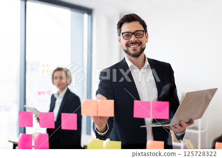 Young adult male and female business professionals collaborating with sticky notes and technology in professional workplace environment, showcasing teamwork, brainstorming 125728322