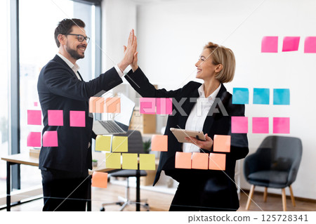 Male and female business professionals collaborate in office using sticky notes on glass board. Team displays successful planning energy highlighting cooperation, positivity achieving corporate goals. 125728341
