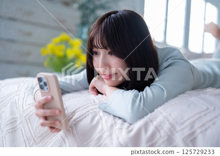 Smiling young woman relaxing while lying face down on her bed and looking at her smartphone screen 125729233