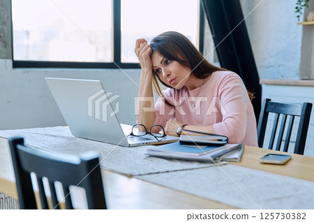 Middle aged sad stressed woman sitting at home at table with computer 125730382