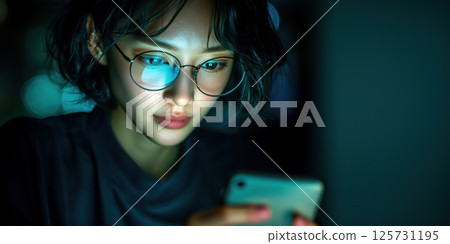 Asian woman with glasses using smartphone at night, focused expression, blue light reflection 125731195
