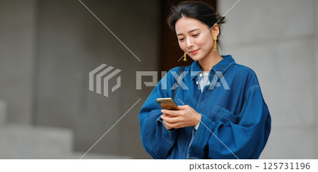 Asian woman using smartphone outdoors, smiling, casual blue shirt, modern city background 125731196