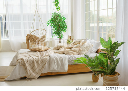 Comfortable bed with lots of soft decorative beige pillows in light sunny bedroom with large window 125732444