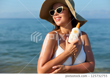 Woman applying sunscreen on her arm at the beach, enjoying a sunny day while wearing a  hat and sunglasses, emphasizing the importance of skin protection. 125732755