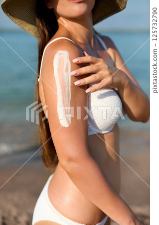 Young woman wearing a white bikini enjoys a sunny day at the beach, relaxing by the water, embodying a carefree summer lifestyle and healthy living. 125732790