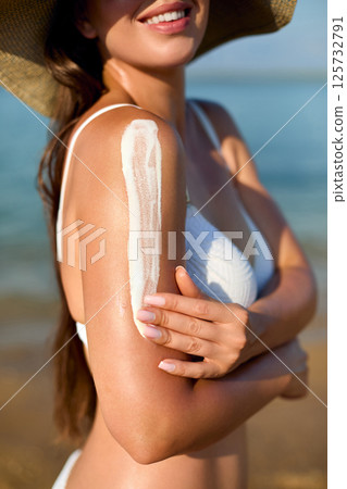 Woman applying sunscreen on her arm while at the beach, enjoying the summer sun and ensuring skin protection from harmful UV rays, a skincare routine. Woman applying sunscreen on her arm while at the beach, enjoying the summer sun and ensuring skin protection from harmful UV rays, a skincare routine. 125732791