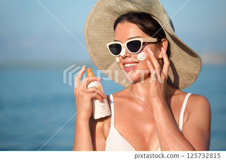 Beautiful young woman with sun cream on face. Girl holding sunscreen bottle on the beach and applying moisturizing lotion on skin.Skin care concept. Sun protection. Suntan Beautiful young woman with sun cream on face. Girl holding sunscreen bottle on the beach and applying moisturizing lotion on skin.Skin care concept. Sun protection. Suntan 125732815