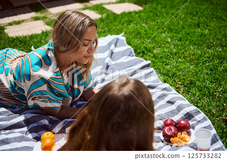 Two friends share a joyful afternoon picnic outdoors, savoring fresh fruit and enjoying sunny weather while lounging on a striped blanket on lush green grass. Two friends share a joyful afternoon picnic outdoors, savoring fresh fruit and enjoying sunny weather while lounging on a striped blanket on lush green grass. 125733282