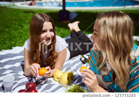 Two women are seated on a blanket outdoors, enjoying fresh fruits during a sunny and relaxing day by the pool. The scene radiates friendship and leisure. 125733283