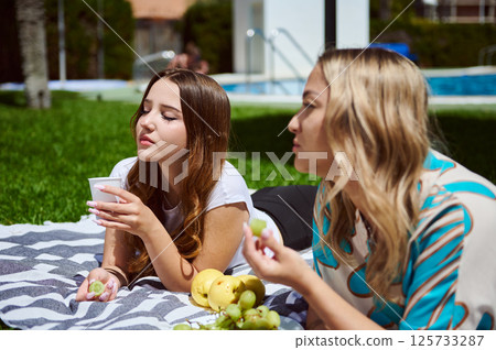 Friends relax on a picnic with fresh fruit and drinks by the poolside. A serene moment conveying friendship, leisure, and happiness under the warm sunlight. 125733287