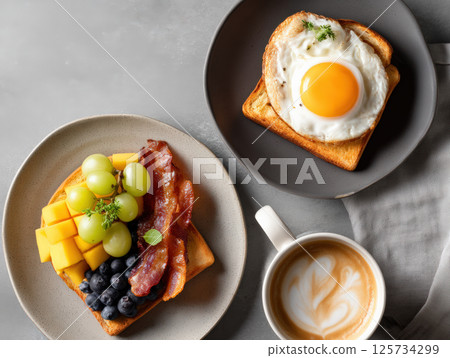 Breakfast toast with fried egg and fresh fruit including grapes, blueberries, cheese, and crispy bacon served with cup of coffee on gray table 125734299
