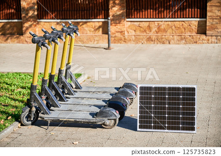 Row of five electric scooters with yellow handlebars parked on sidewalk next to patch of grass. Solar panel placed nearby, indicating eco-friendly sustainable charging solution for scooters. 125735823