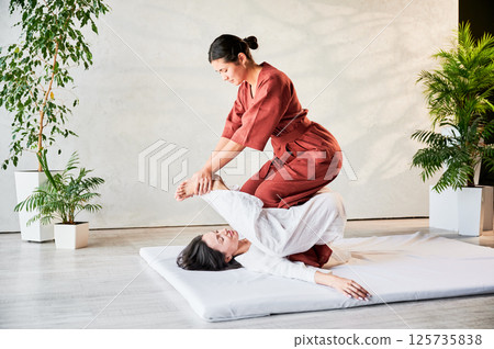 Masseuse making thai yoga massage. Woman therapist doing traditional massage treatment, stretching female patient's body. Client lying on mat, enjoying hand massage in calm atmosphere. Masseuse making thai yoga massage. Woman therapist doing traditional massage treatment, stretching female patient's body. Client lying on mat, enjoying hand massage in calm atmosphere. 125735838