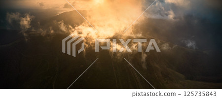 Stunning aerial view of mountain landscape at sunrise, with sun's golden rays piercing through mist and clouds, casting magical glow over peaks, creating serene and ethereal atmosphere. Aerial view. Stunning aerial view of mountain landscape at sunrise, with sun's golden rays piercing through mist and clouds, casting magical glow over peaks, creating serene and ethereal atmosphere. Aerial view. 125735843