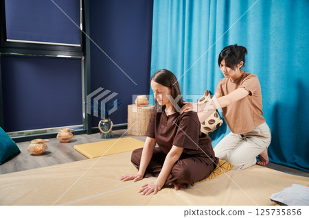 Masseuse making thai yoga massage. Woman therapist doing traditional massage treatment for female patient indoors. Client sitting on mat, meditating, enjoying hand massage in calm atmosphere. Masseuse making thai yoga massage. Woman therapist doing traditional massage treatment for female patient indoors. Client sitting on mat, meditating, enjoying hand massage in calm atmosphere. 125735856