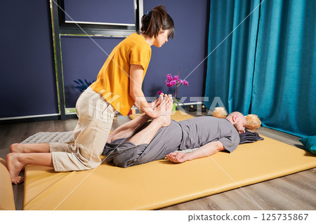 Masseuse making thai yoga massage. Woman therapist doing traditional massage treatment, stretching male patient's body. Bald client lying on mat, enjoying hand massage in calm atmosphere. Masseuse making thai yoga massage. Woman therapist doing traditional massage treatment, stretching male patient's body. Bald client lying on mat, enjoying hand massage in calm atmosphere. 125735867