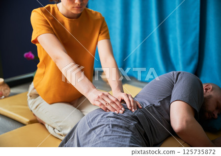 Masseuse making thai yoga massage. Cropped view of woman therapist doing traditional massage treatment for male patient indoors. Client lying on mat, enjoying hand massage in calm atmosphere. Masseuse making thai yoga massage. Cropped view of woman therapist doing traditional massage treatment for male patient indoors. Client lying on mat, enjoying hand massage in calm atmosphere. 125735872