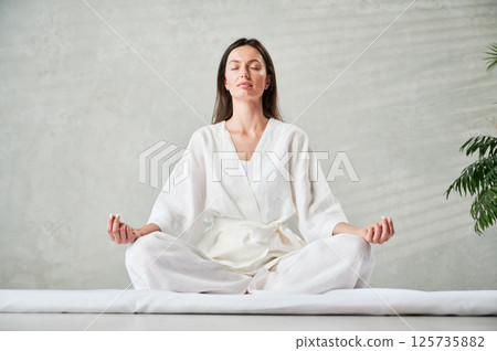Beautiful woman in white dress sitting on mat in lotus position before Thai massage, practicing yoga. Beautiful woman in white dress sitting on mat in lotus position before Thai massage, practicing yoga. 125735882