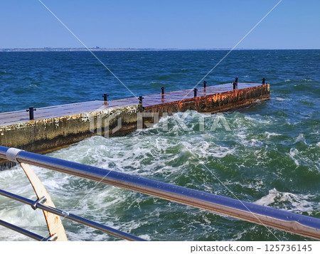 Stormy sea. Large sea waves crashing and hitting against old concrete pier on sunny summer day. High waves with white foam. Big waves breaking, spreading on cement pier. Splashes bursts and splashing Stormy sea. Large sea waves crashing and hitting against old concrete pier on sunny summer day. High waves with white foam. Big waves breaking, spreading on cement pier. Splashes bursts and splashing 125736145