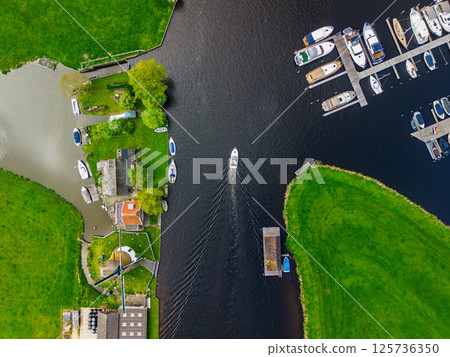 A top-down view of a marina with multiple boats docked along wooden piers, a boat cruising through a narrow waterway surrounded by green parks and natural grassy areas. A top-down view of a marina with multiple boats docked along wooden piers, a boat cruising through a narrow waterway surrounded by green parks and natural grassy areas. 125736350