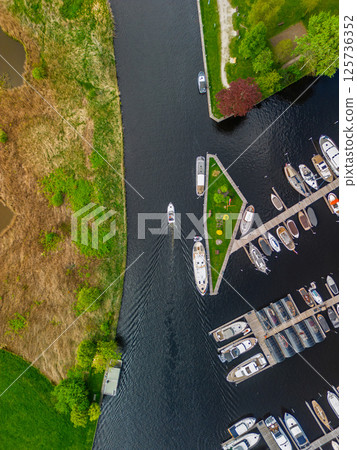 A top-down view of a marina with multiple boats docked along wooden piers, a boat cruising through a narrow waterway surrounded by green parks and natural grassy areas. 125736352