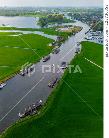 Aerial view of a peaceful Dutch landscape with green fields, a canal lined with houseboats, a traditional windmill, and a marina in the distance under a cloudy sky. Aerial view of a peaceful Dutch landscape with green fields, a canal lined with houseboats, a traditional windmill, and a marina in the distance under a cloudy sky. 125736355