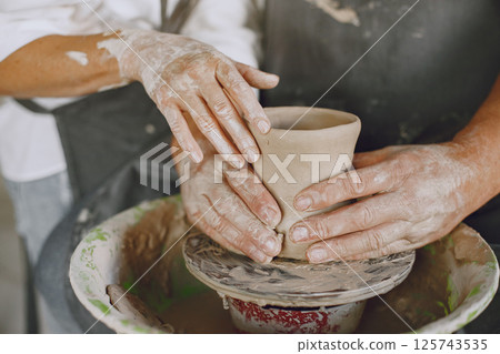 Mutual creative work. Adult elegant couple in casual clothes and aprons. People creating a bowl on a pottery wheel in a clay studio. Mutual creative work. Adult elegant couple in casual clothes and aprons. People creating a bowl on a pottery wheel in a clay studio. 125743535