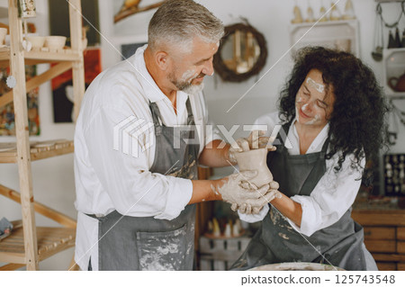 Mutual creative work. Adult elegant couple in casual clothes and aprons. People creating a bowl on a pottery wheel in a clay studio. Mutual creative work. Adult elegant couple in casual clothes and aprons. People creating a bowl on a pottery wheel in a clay studio. 125743548