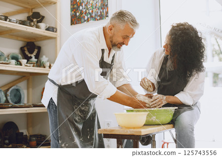 Mutual creative work. Adult elegant couple in casual clothes and aprons. People creating a bowl on a pottery wheel in a clay studio. 125743556
