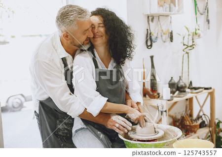 Mutual creative work. Adult elegant couple in casual clothes and aprons. People creating a bowl on a pottery wheel in a clay studio. Mutual creative work. Adult elegant couple in casual clothes and aprons. People creating a bowl on a pottery wheel in a clay studio. 125743557