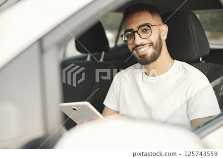 Handsome indian young man looking at camera while sitting in the car. Man wearing white t-shirt and glasses. Male driver sitting on car and hand holding smartphone to use online application. 125743559