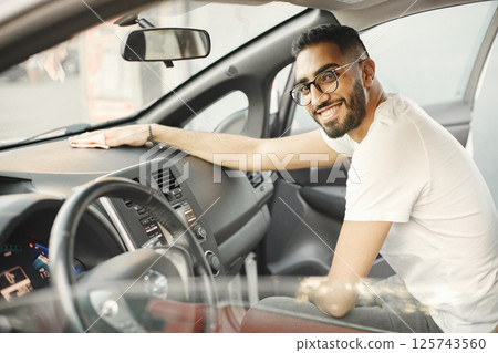 Young man in glasses polishing inside his car with a rug. Man wearing white t-shirt. Indian man wiping his vehicle. 125743560