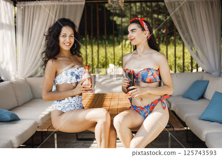 Two female friends in a swimwear sitting in bungalow. Girls wearing blue and red swimwear. Girls posing for a photo in a bungalow and holding a cocktails. 125743593