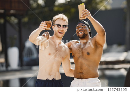 Caucasian and black american boys relaxing near swimming pool. Boys wearing white and black swimwear and a sunglasses. Men holding a cocktails. Caucasian and black american boys relaxing near swimming pool. Boys wearing white and black swimwear and a sunglasses. Men holding a cocktails. 125743595