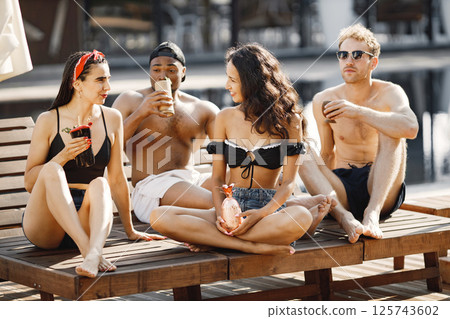 Two young girls and two theirs male friends relaxing on a sunbeds near swimming pool. Girls wearing black swimwear and red headband. Black american boy, caucasian boy and girls drinking cocktails. 125743602
