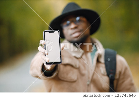Cropped portrait of black male traveler showing blank copy screen cell phone for your information or advertisement. Selective focus. Man standing outside and wearing a brown jacket, glasses and black 125743640