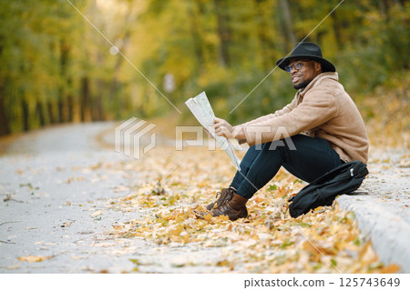 Young black man sitting on road and looking at map. Male traveler feeling lost, traveling alone by autostop. Man wearing brown jacket, black hat and backpack. 125743649