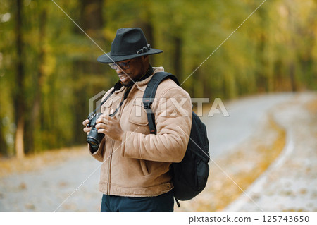 Young black man standing on road in forest with a camera. Male photographer walking in a forest. Man wearing brown jacket, black hat and backpack. 125743650