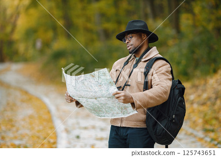 Young black man hitchhiking on road and looking at map. Male traveler feeling lost, traveling alone by autostop. Man wearing brown jacket, black hat and backpack. 125743651