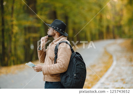 Young black man hitchhiking on road and holding a map. Male traveler feeling lost, traveling alone by autostop. Man wearing brown jacket, black hat and backpack. 125743652