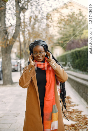 Young black woman with a long locs hairstyles standing outside. Woman wearing brown coat, orange scarf and black hat. Woman in glasses using a white earphones. Young black woman with a long locs hairstyles standing outside. Woman wearing brown coat, orange scarf and black hat. Woman in glasses using a white earphones. 125743659