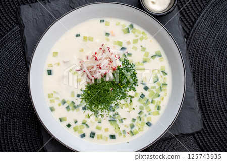 Cold okroshka summer soup nestled in white ceramic bowl, topped with chopped dill, chives, grated radish, accompanied by small sour cream side dish on dark slate serving plate Cold okroshka summer soup nestled in white ceramic bowl, topped with chopped dill, chives, grated radish, accompanied by small sour cream side dish on dark slate serving plate 125743935