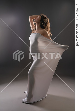 Beautiful pregnant woman posing gracefully in a flowing white garment Beautiful pregnant woman posing gracefully in a flowing white garment 125746704