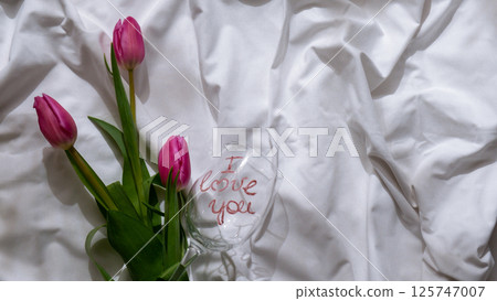 I LOVE YOU message on wine glass with fresh tulips on white bed with trendy deep shadows. Spring flowers in pink violet colors. Romantic postcard wallpaper design 125747007
