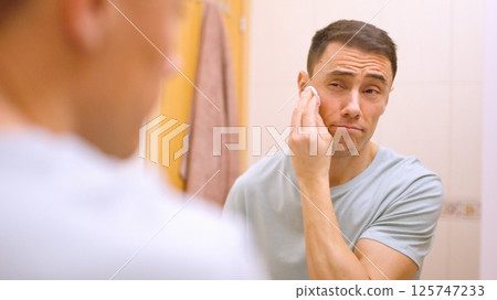 Man cleansing face skin with cotton pad in bathroom Man cleansing face skin with cotton pad in bathroom 125747233