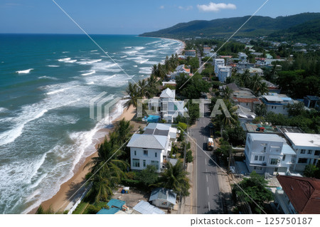 Aerial view of coastal town with scenic beach and road lined with palm trees and modern houses, set against backdrop of lush green hills and clear blue sky 125750187